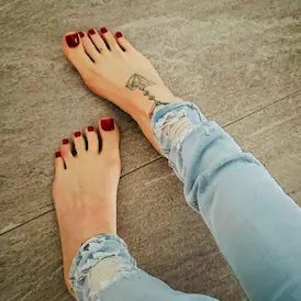 Barefoot with red toenails and her foot tattoo in ripped jeans on a grey floor - Foot Fetish Zurich